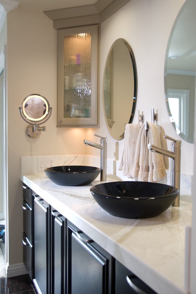 Remodeled dual bathroom vanity with dark cabinets, white stone countertops, black bowl sinks with modern brushed-nickel faucets, and a round mirror on the wall above each sink. Gray glass-front cabinet in the far corner wall.
