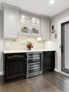 Home bar area with wood floors, black cabinets below and white cabinets above. White stone countertop and white stone backsplash. Glass-front cabinets in middle of uppers. Wine fridge in middle of lower cabinets.