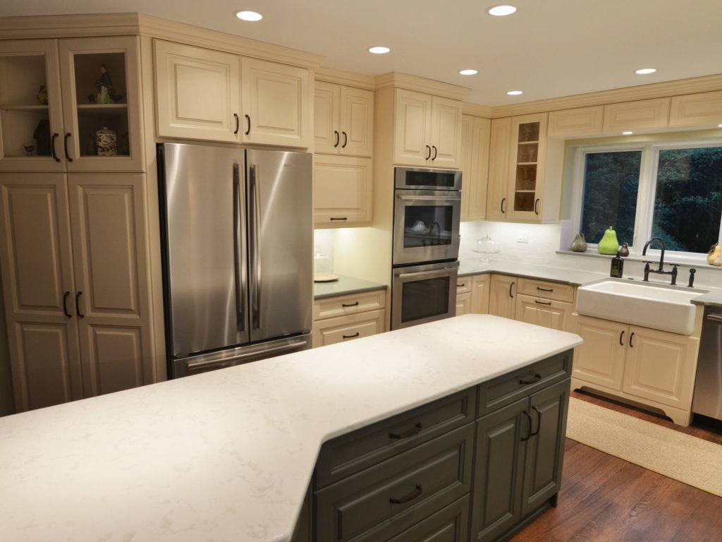 Kitchen renovation with cream cabinets, L-shaped island with green cabinets, white stone countertops, recessed lighting, stainless steel appliances, white farmhouse sink, and wood floors.