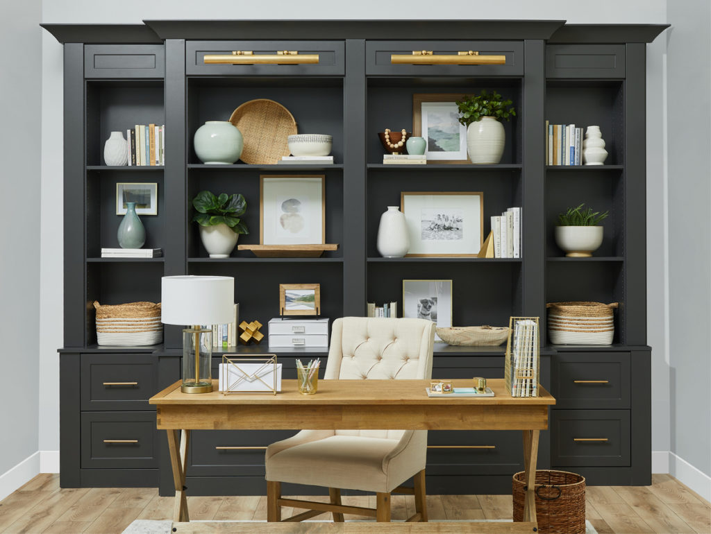 Remodeled home office with built-in black shelving and drawers on far wall behind a light wood desk. Light wood floors. Light gray walls. Gold hardware and accent pieces.