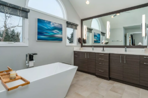 Remodeled contemporary bathroom with white soaking tub, dark cabinets, white tile floors, natural light from 3 windows on left wall. 
