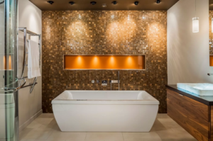 Modern glamour style bathroom remodel with large white freestanding tub, gold tile accent wall behind the tub, with built-in shelf with yellow lighting. Large-format tan tile floor, woodgrain modern cabinetry.