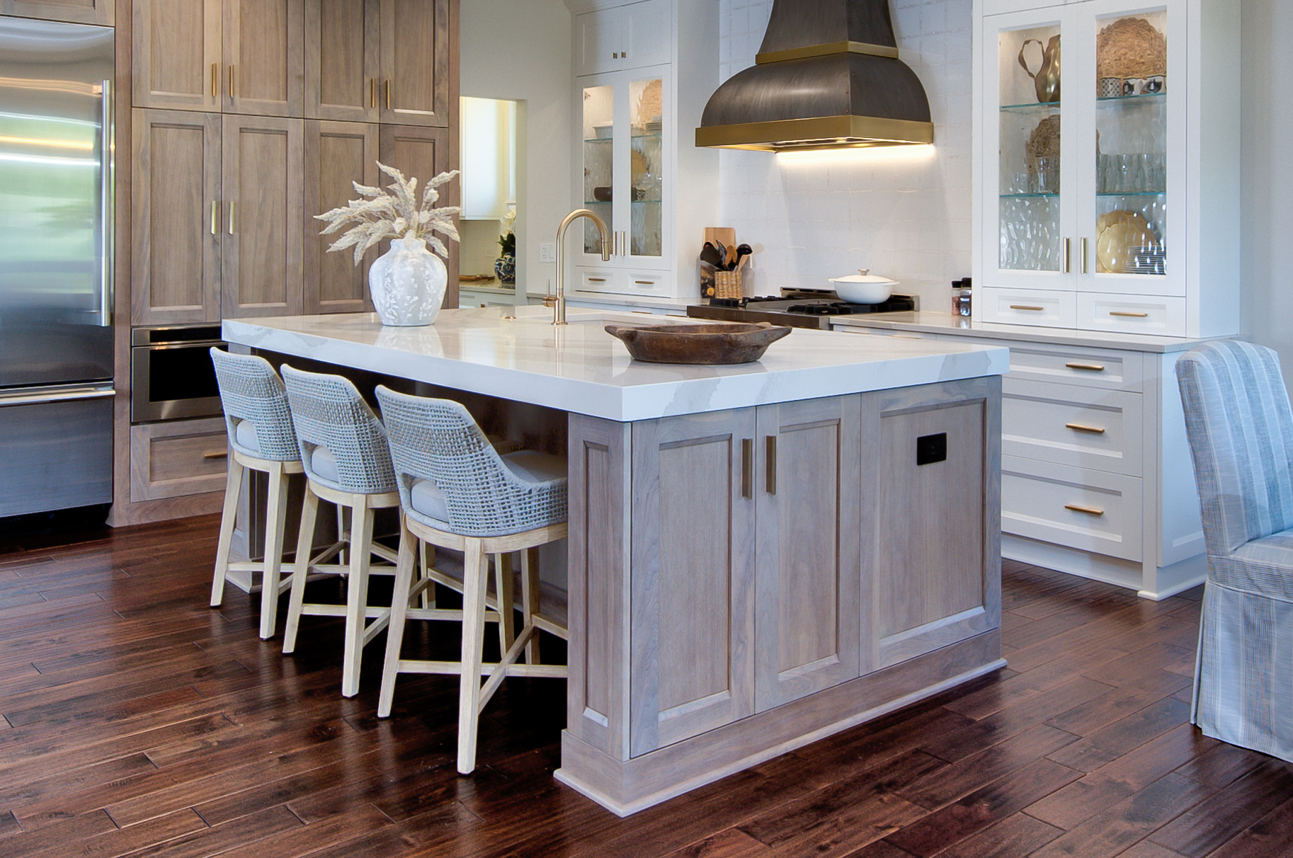 Remodeled kitchen with large island with light woodgrain cabinets and thick white stone countertop and three stools. White cabinets against back wall. Wood floors.