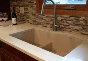 Recently installed deep double sink with surrounding white stone countertop, modern brushed nickel faucet, brown-tones glass tile backsplash.