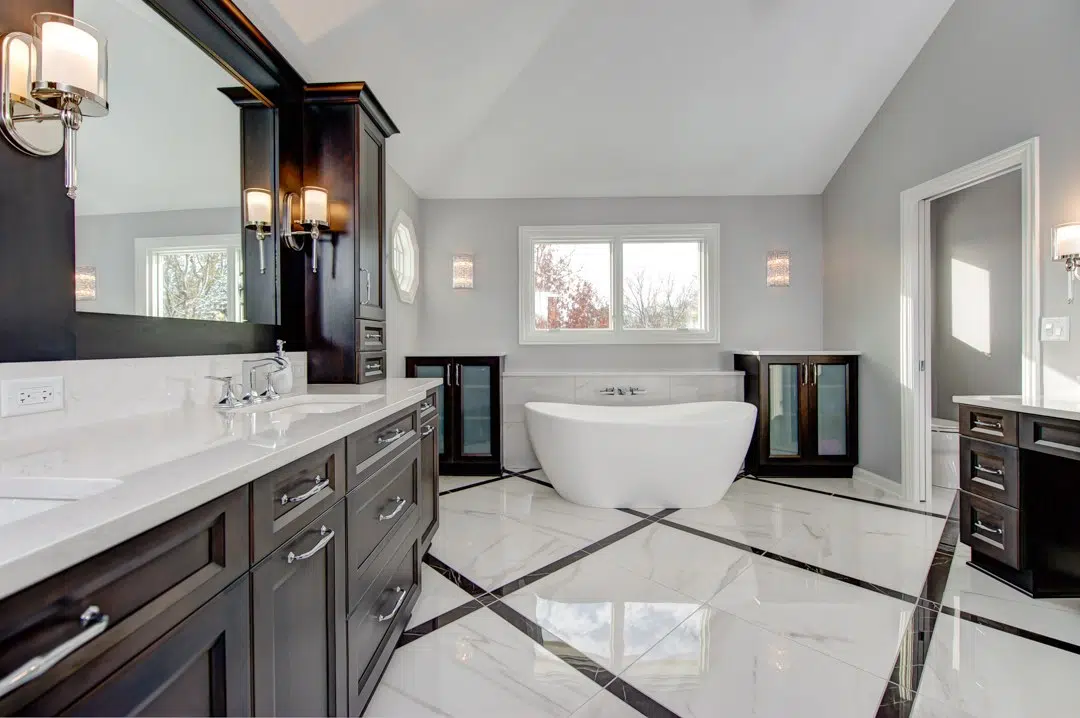 Primary bathroom remodel with large white tile flooring, standalone white soaking tub at far end of room under a small sliding window, dark woodgrain dual vanity to the left with white stone countertop.