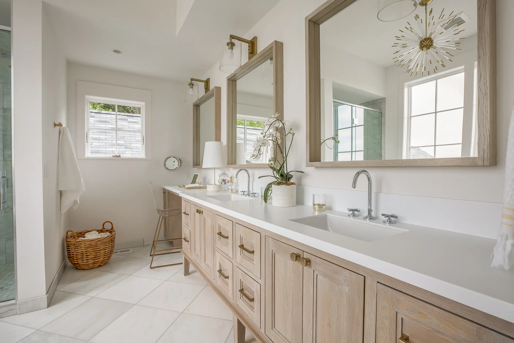 Remodeled bathroom with light woodgrain double vanity with white stone countertop, white tile floors, white walls, three large mirrors with matching light wood frames on wall above vanity. Small window in wall at far end.