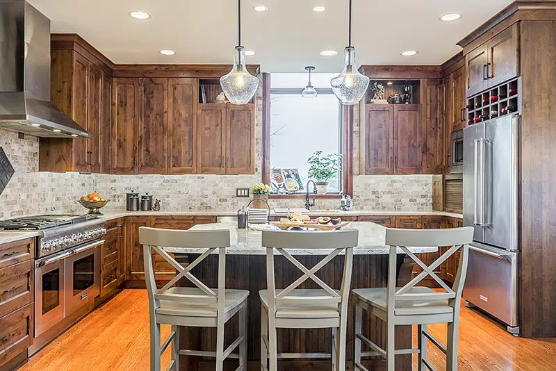 The Kitchen Master remodeled kitchen with pendant lights, hooded vent, white brick backsplash, and woodgrain shaker cabinets. Island in the middle with stool seating. Stone countertops. Wood floors.