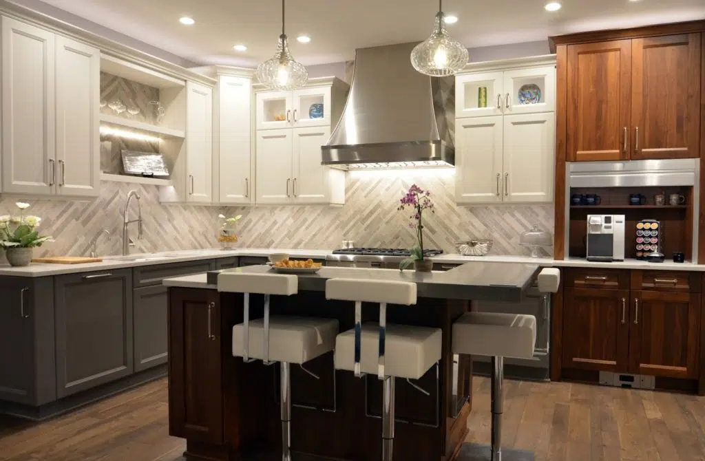 Kitchen remodel with wood floors, gray lower cabinets and white upper cabinets, white stone countertops, herringbone glass tile backsplash, and small square island with white stool seating.