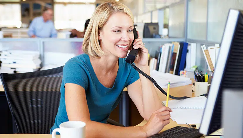 Woman sitting at desk looking at computer screen holding phone and pencil smiling