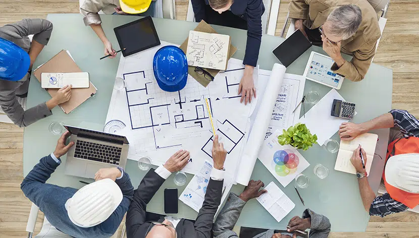 Eight men sitting around a table with architectural plans rulers laptops and waters between them