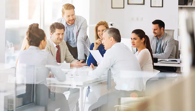 Seven people sitting at a table talking with papers and glasses of water between them