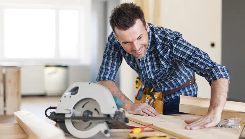 carpenter holding wood onto a table beside a saw wearing a tool belt