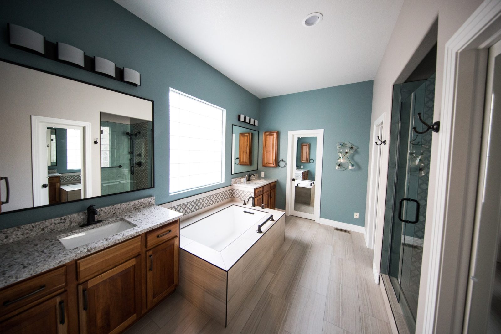 Custom bathroom remodel with blue walls, large picture window over separate soaking tub, with vanity on either side featuring woodgrain cabinets and stone countertops. Light wood-look tile flooring.