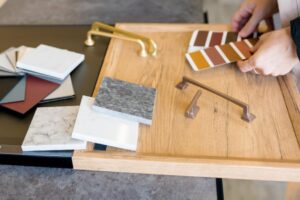 A person's hands holding several paint color samples over a small wooden workdesk, alongside counter material samples and different styles of cabinet handles.