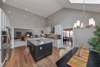 Modern, open kitchen remodel with vaulted ceiling, large square island, wood floors, dining area in foreground, light gray walls.